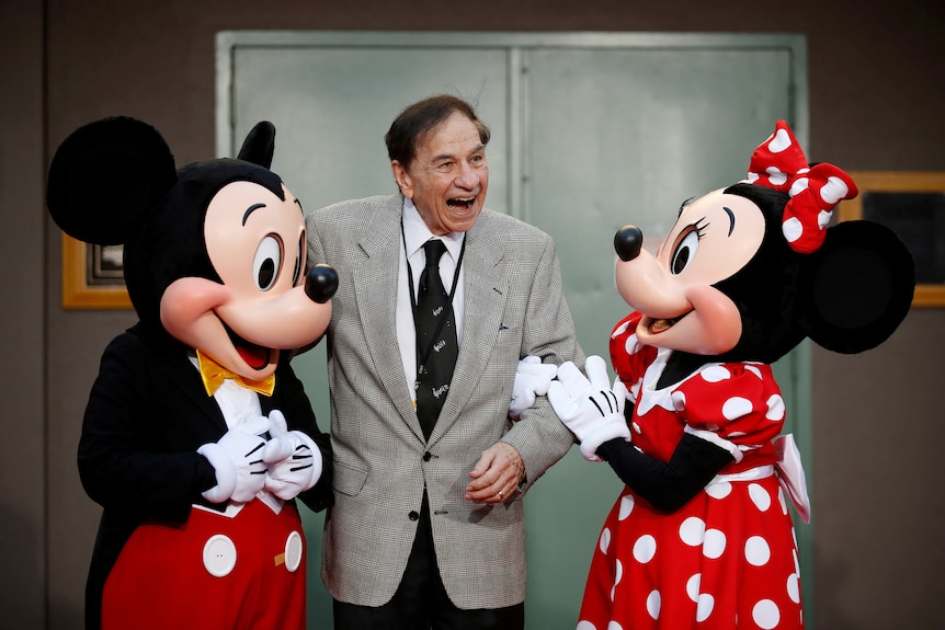 A man smiles while standing next to cartoon mascots of two mice.