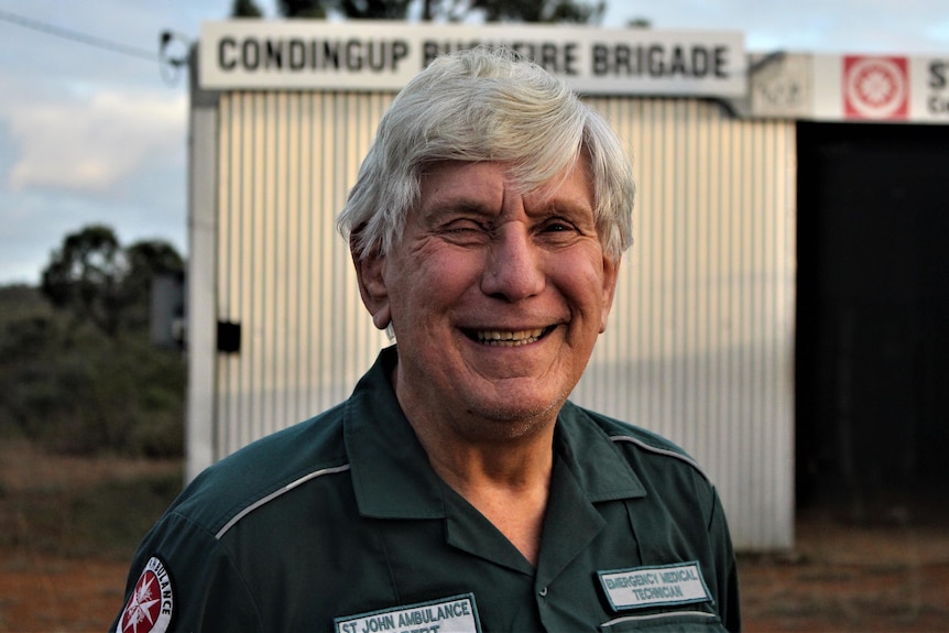 A laughing grey-haired man stands in front of the tiny ambulance shed.
