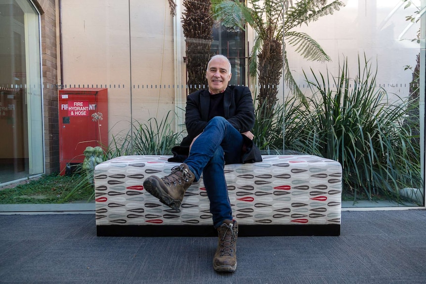 A man in blue jeans sits on a couch in front of a glass window.