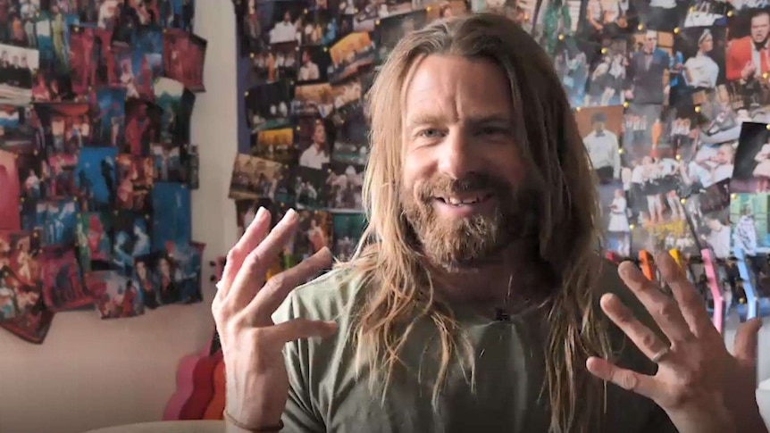 A man with long hair and a beard chats in front of a wall mounted with guitars and boards covered with photos of school students