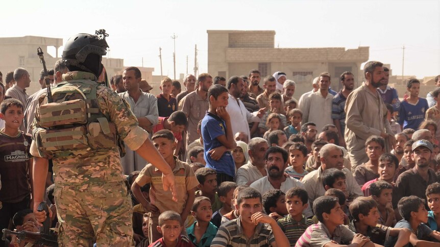 Displaced people sit in a large group on the ground, as a soldier stands before them.