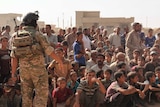 Displaced people sit in a large group on the ground, as a soldier stands before them.