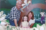 A husband and wife standing behind their daughter and in front of a colourful children's room background 