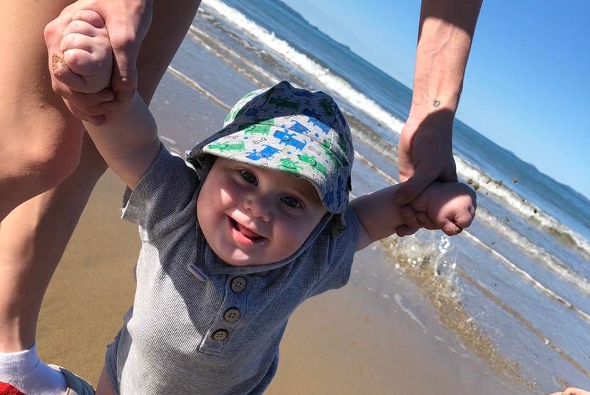 Young baby held by his arms walks on sand at the beach.