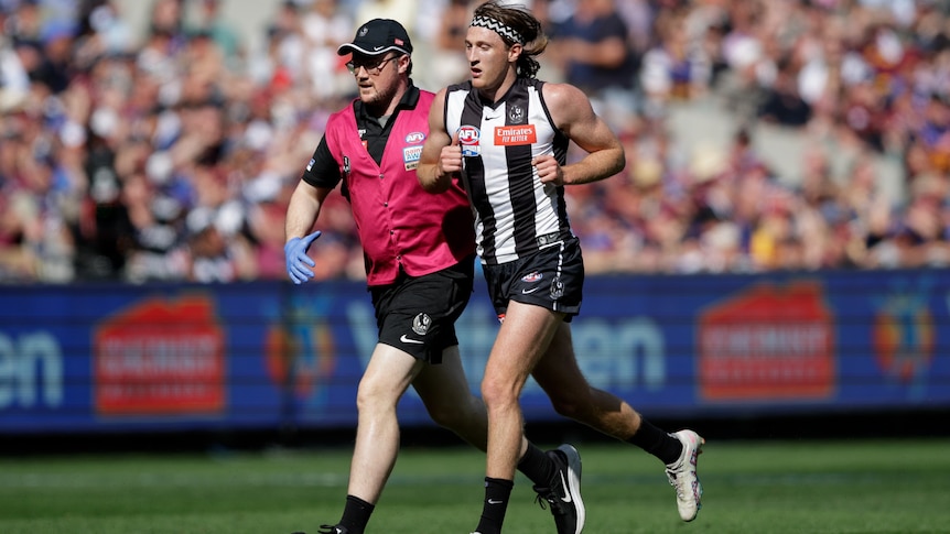A Collingwood AFL player runs from the field with the assistant of a trainer during the 2023 grand final at the MCG.