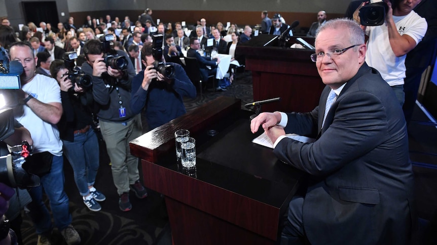 Morrison sits behind a lectern in front of a wall of people with cameras.