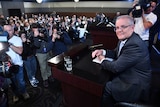 Morrison sits behind a lectern in front of a wall of people with cameras.