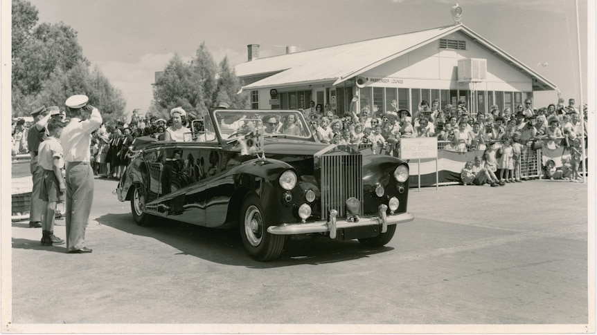 A black and white photo of queen elizabeth in the passenger seat of the car with police and crowds surrounding her