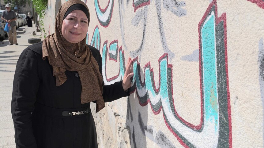 A woman wearing a religious head covering stands in front of a mural that contains Arabic text