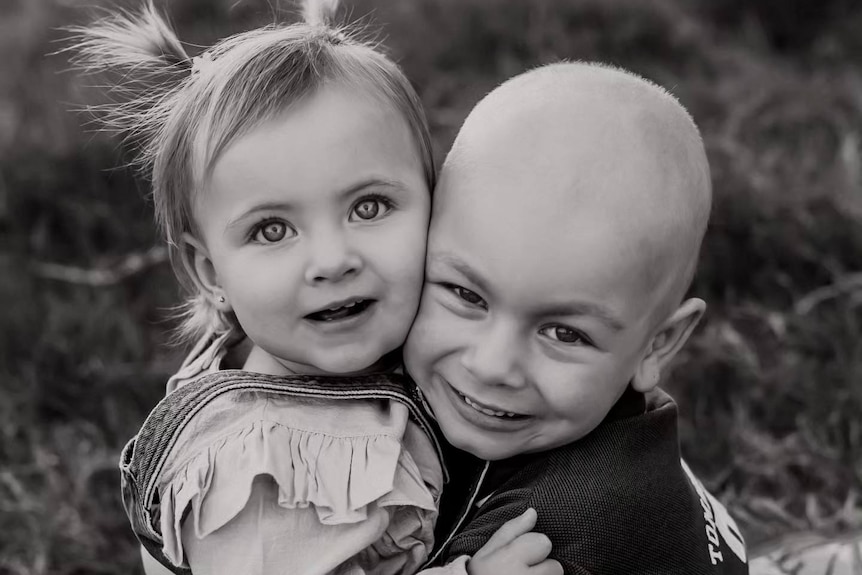 A black-and-white photo of a young boy with a shaved head cuddling his toddler sister.