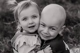 A black-and-white photo of a young boy with a shaved head cuddling his toddler sister.