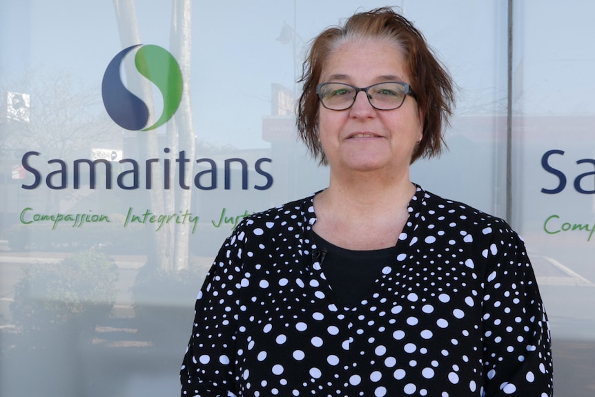 A lady wearing glasses and a black and white polka-dot shirt stands in front of a Samaritans logo on a shop front window.