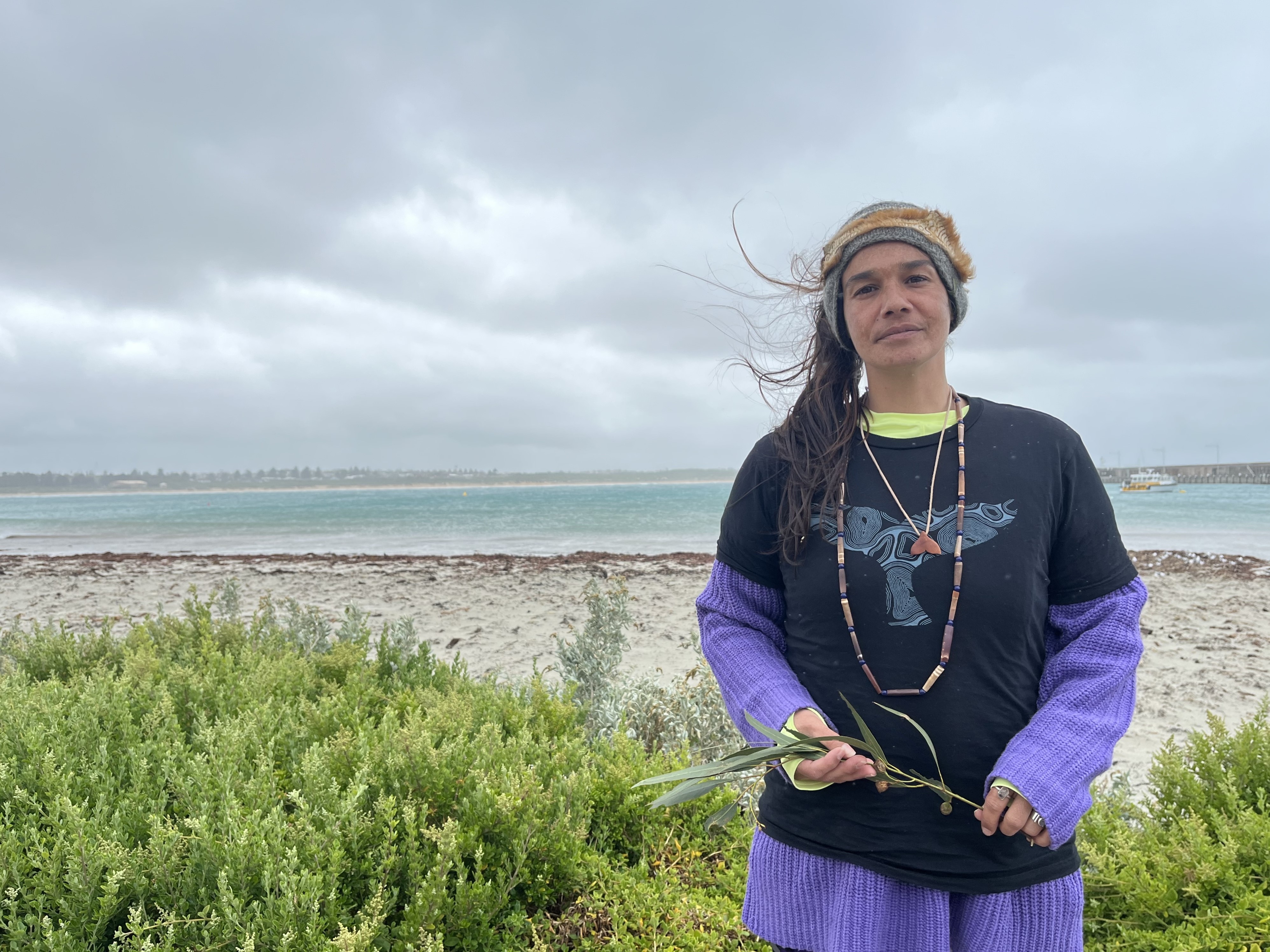 A woman stands in front of the ocean holding a branch from a plant. 