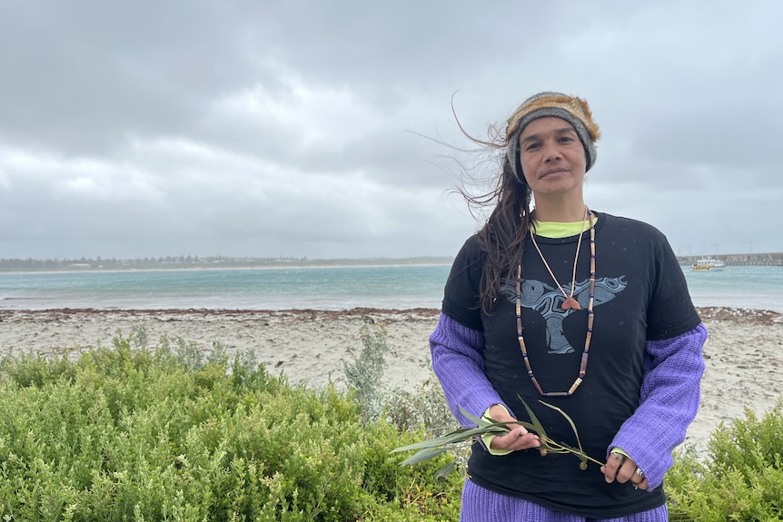 A woman stands in front of the ocean holding a branch from a plant. 