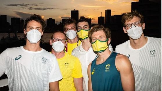 A group of mask-wearing Olympic swimmers pose in front of a city skyline at sunset.