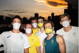 A group of mask-wearing Olympic swimmers pose in front of a city skyline at sunset.