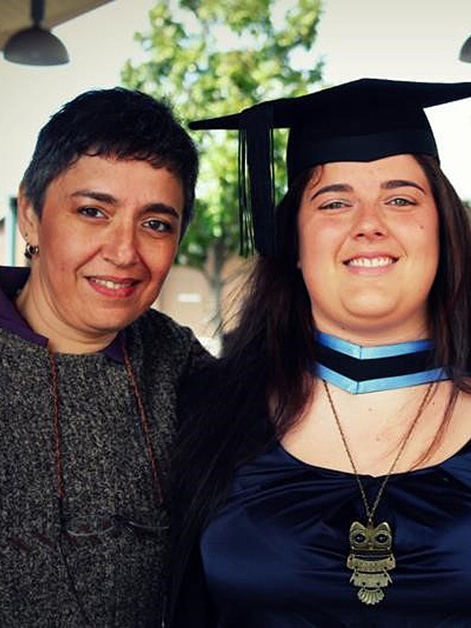 A mother and daughter smile at the camera. The daughter is wearing a mortarboard graduate cap and a necklace of an owl.