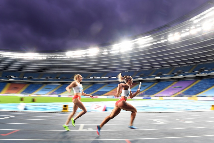 Two women complete a baton hand-off in an athletics relay in an empty stadium