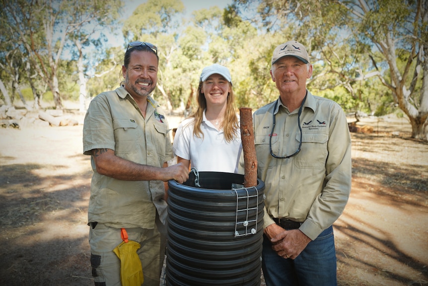 A young girl stands in the middle of two older men, all three smiling and holding a black plastic artificial bird's nest
