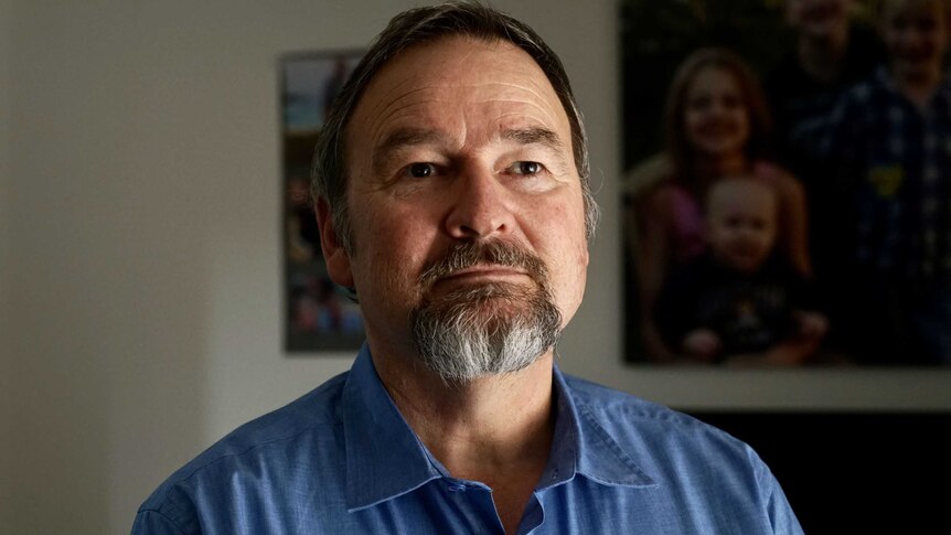 A middle-aged man with a greying beard stands tall, wearing a blue shirt and looking off-camera