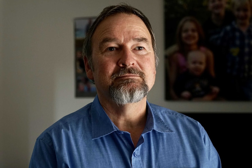 A middle-aged man with a greying beard stands tall, wearing a blue shirt and looking off-camera