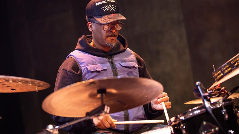 Emanuel Harrold sits comfortably at his drum kit playing, he is wearing a cap, sunglasses and a hoodie with a vest.