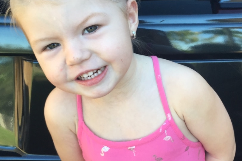 A young child with blonde hair pulled off her face with a pink clip, wearing a pink dress and smiling looking happy