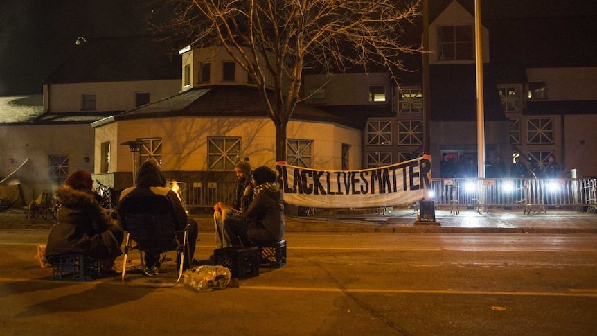 A group sits outside of the police station after five people were shot