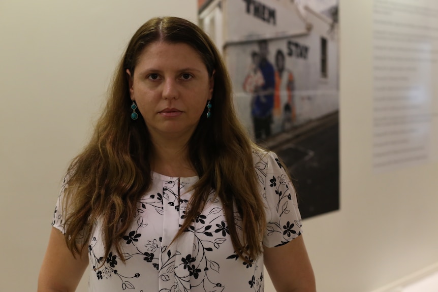 A serious young woman with long brown hair, blue earrings, white and black top, stands in front of a photo of street art.