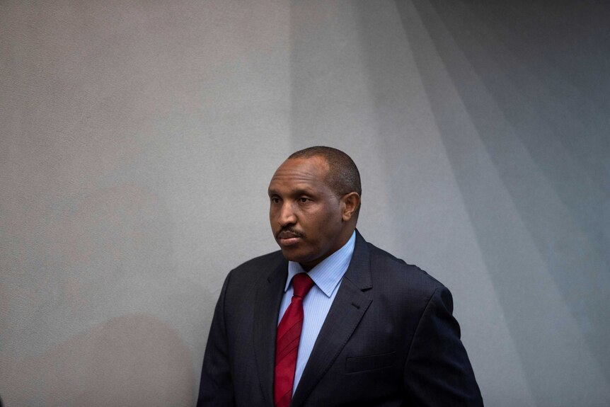 Bosco Ntaganda is pictured against a black wall as he enters a room wearing a dark navy suit with a red tie.