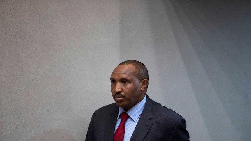 Bosco Ntaganda is pictured against a black wall as he enters a room wearing a dark navy suit with a red tie.