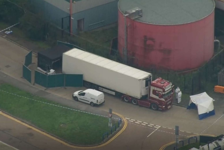 Shot from above of a large truck. It's back is open but cordoned off. People in white jumpsuits mull nearby.