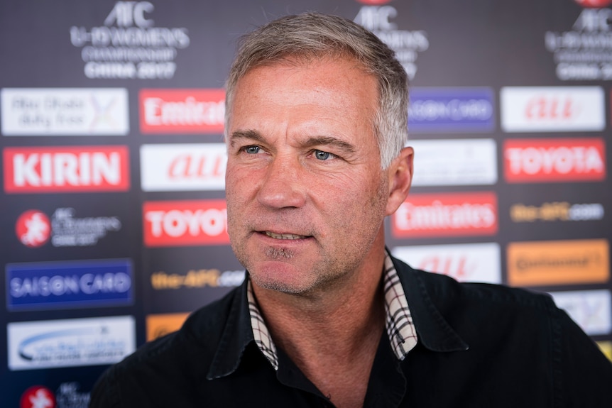 A white man with a dark collared shirt speaks with an advertising board behind him