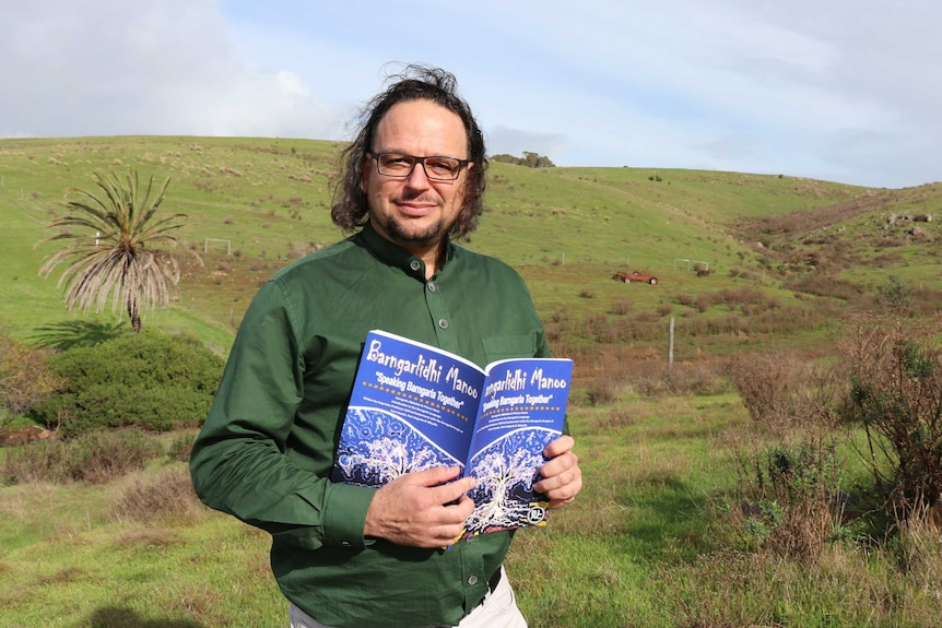 Man in middle of frame holding a book with green paddock in backgound