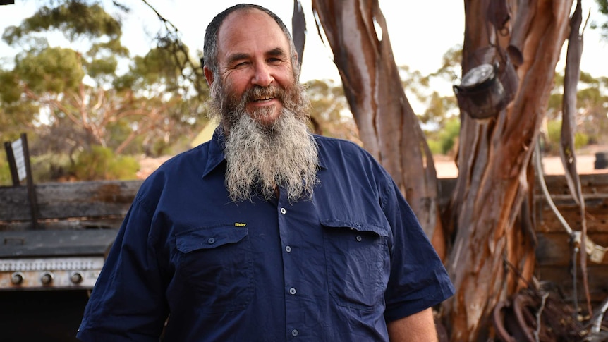 A man with a long beard and wearing a blue shirt smiles at the camera.