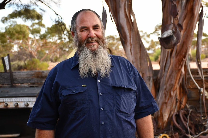 A man with a long beard and wearing a blue shirt smiles at the camera.