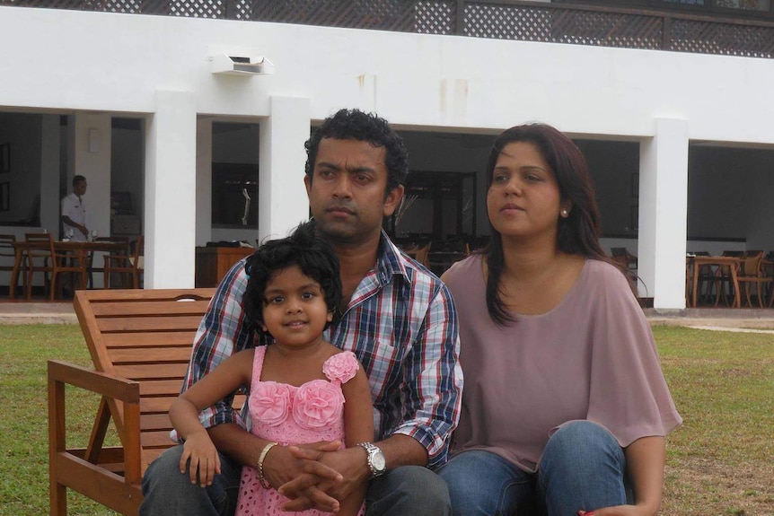 Man and woman sit and look off in the distance as girl in pink dress and short hair smiles at the camera.