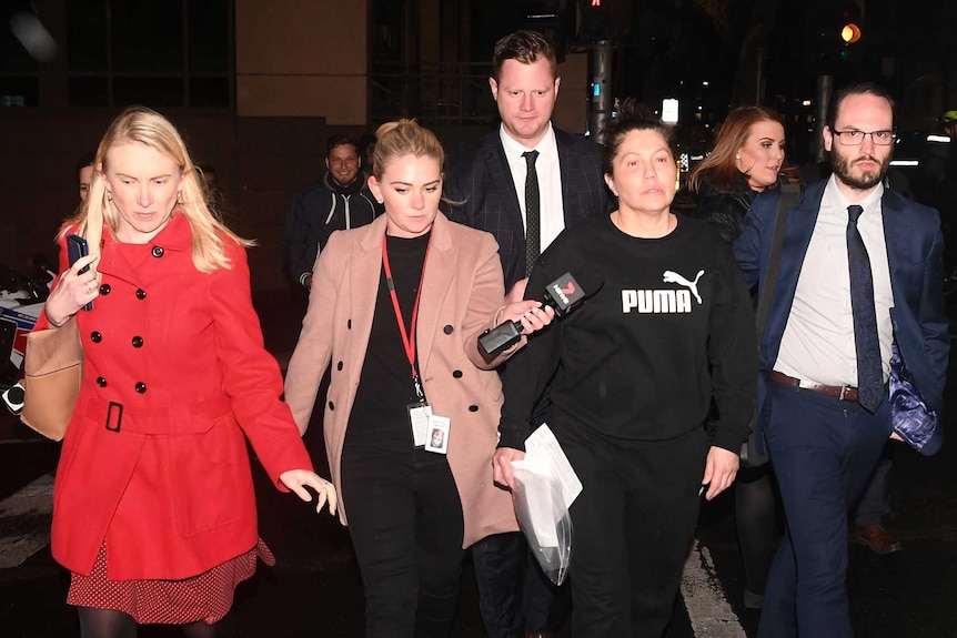 Roberta Williams walks away from court in the dark, followed by men in suits, while two female reporters ask her questions.