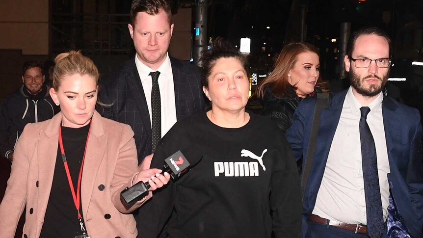 Roberta Williams walks away from court in the dark, followed by men in suits, while two female reporters ask her questions.