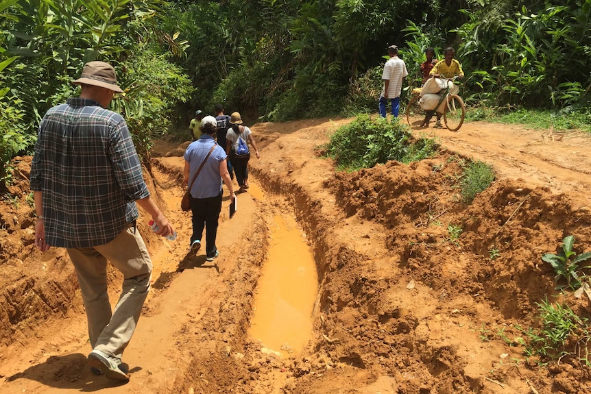 People walking next to deep trench in muddy road while two people wheel bike on bank opposite.