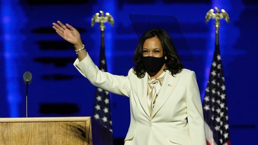Kamala Harris, dressed all in white, smiles from behind a face mask as she waves to a crowd