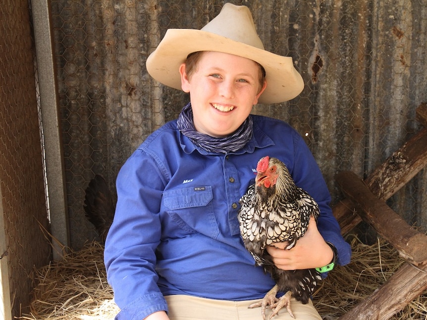 A young boy in a hat and blue shirt, sitting in a chicken coop holding a chicken.