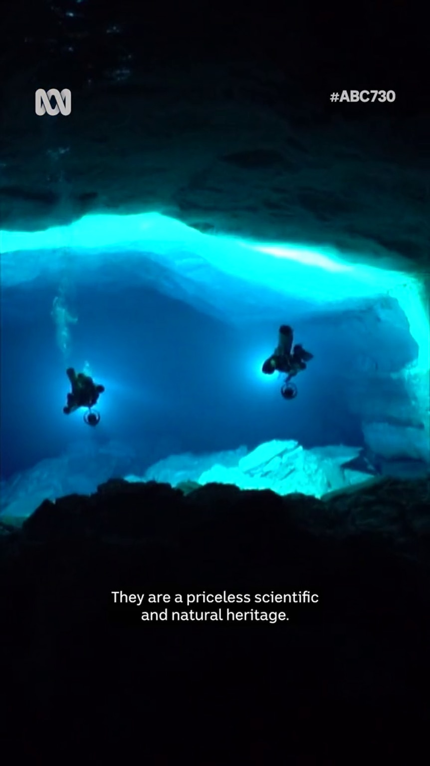 An underwater cave filled with blue-coloured water hosts two dark figures in diving gear