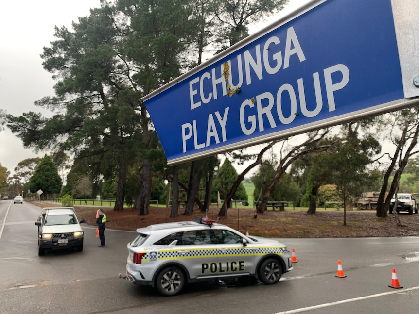A sign saying ECHUNGA PLAY GROUP above a police car and a police officer speaking to a ute