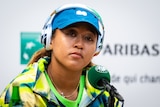 Tennis player Naomi Osaka sits behind a microphone while wearing a cap and headphones during a press conference.