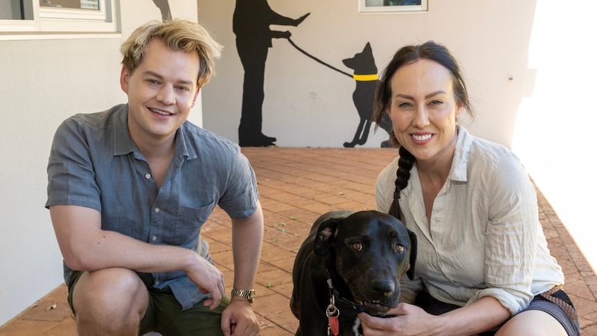 Laura Vissaritis and Joel Creasey out the front of a dog rescue shelter with a black dog in between them