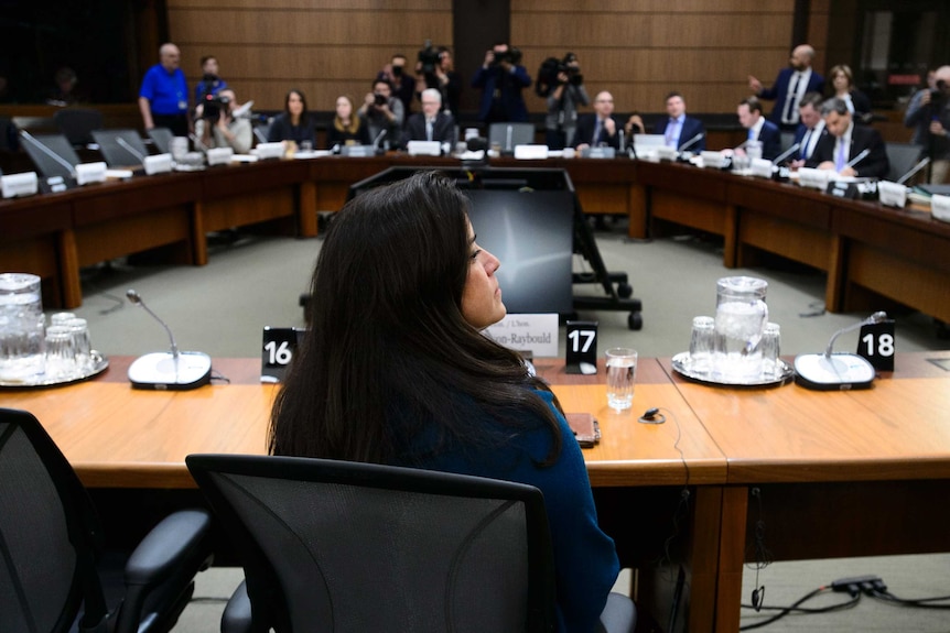 A women sits in front of a room full of people on a round table.