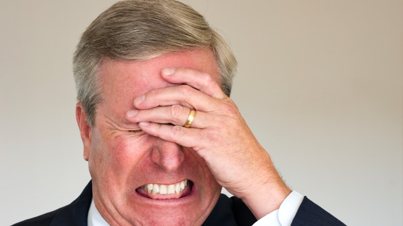 A man with a red face holding his hand to his forehead looking very angry and stressed.