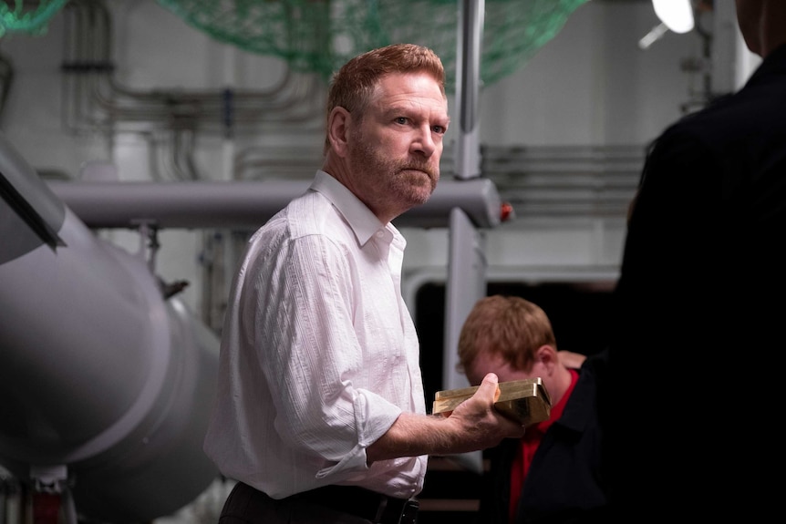 Kenneth Branagh in a white shirt holding bars of gold in the film Tenet