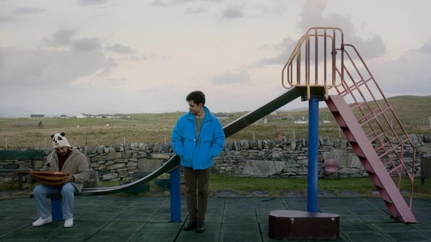 A Syrian man in a bright blue jacket and an Afghan man in a panda hat sit on a metal slide in front of an empty field